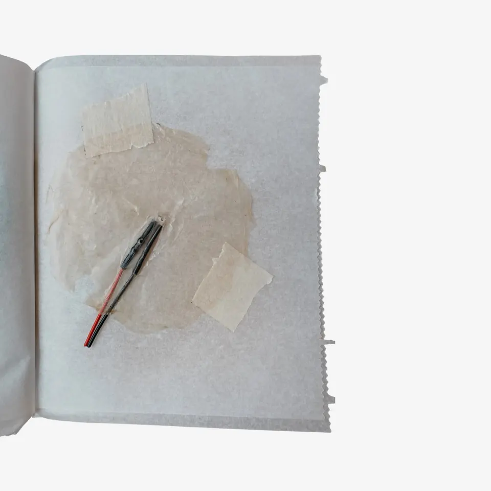 A booklet page with a thin, almost clear sheet and a slim component with red and black wires.