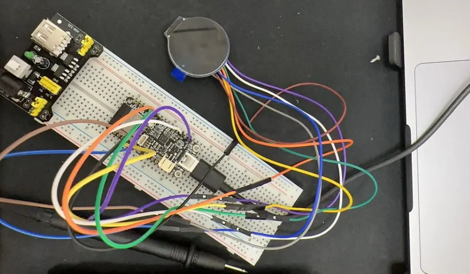 A breadboard wired with colourful jumper cables, a coin battery holder and a small board, on a dark desk.