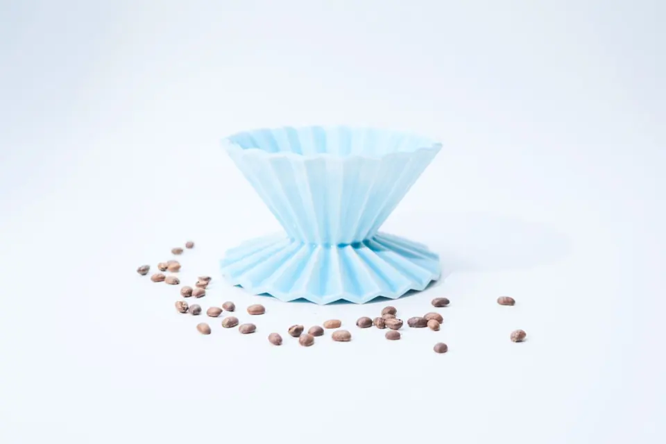 A pale-blue ceramic coffee dripper with pleated, tutu-like walls on a white ground, with coffee beans scattered around it.