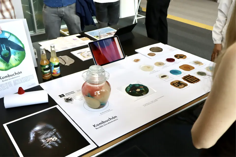 The Kombuchán table at Demo Day: a jar of kombucha, the cicada piece, posters and rows of small samples, with visitors around it.