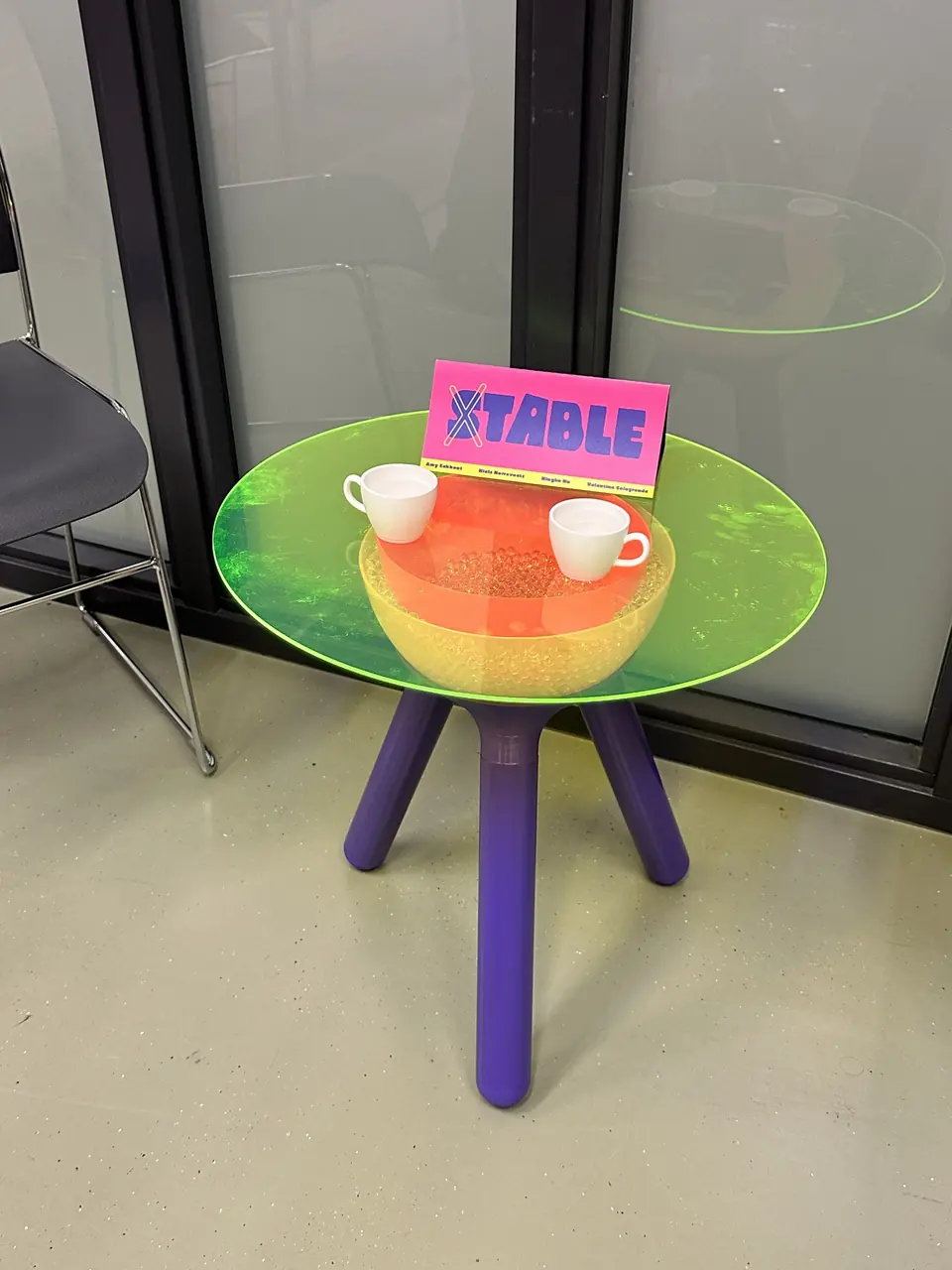 STABLE in the exhibition: a round, glowing acrylic tabletop with cups on purple legs, a pink STABLE sign on top.