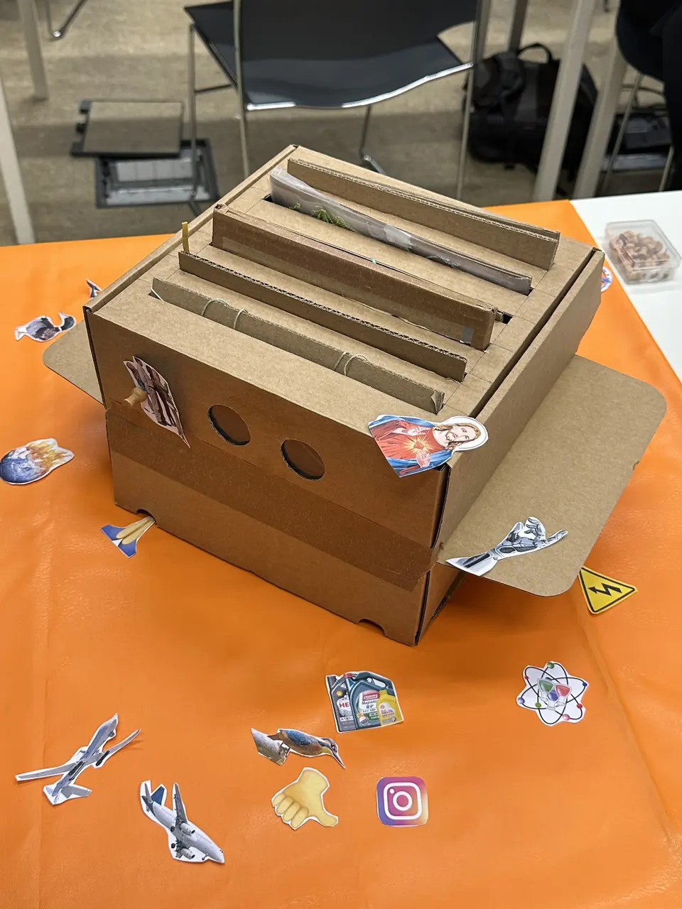 A slotted cardboard box on an orange table, surrounded by cut-out stickers.