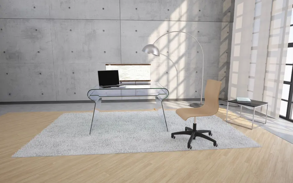 A clear glass desk with a laptop and a wooden chair on a pale wood floor, in a concrete room with sunlit windows.