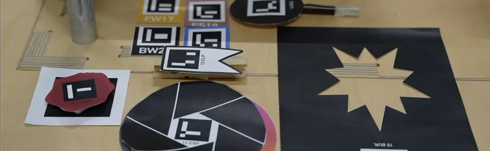 Physical editing tools on a table: an aperture disc, paddles, filter tiles and a red stamp, each marked with an AprilTag.